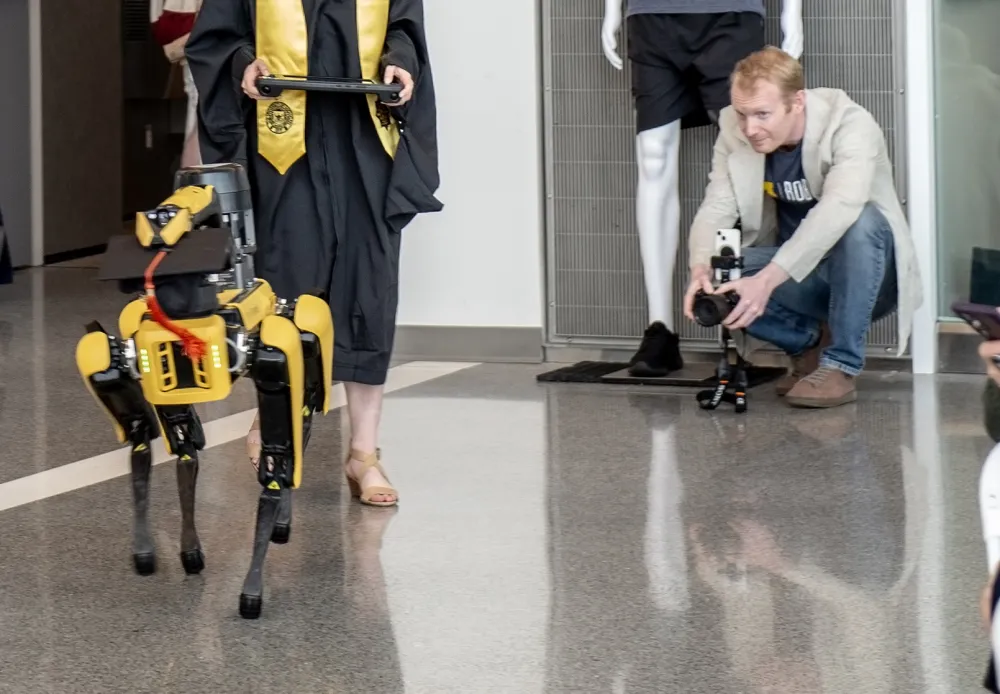 Dan Newman crouches with a camera to film a robotic dog wearing a graduation cap and tassel at a University of Michigan commencement.
